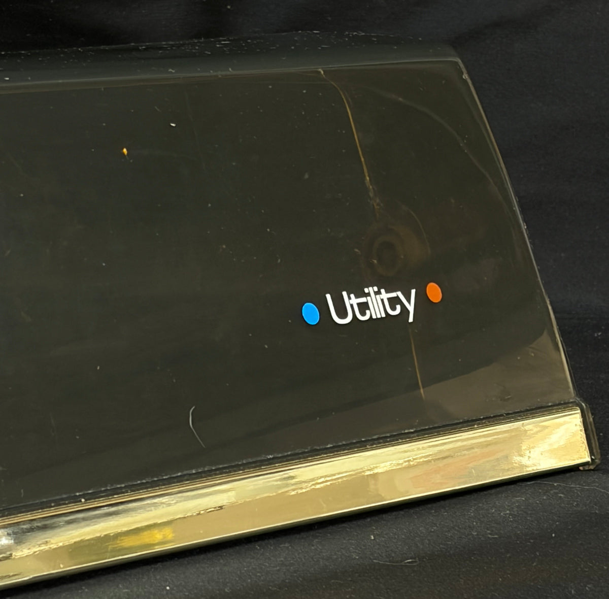 A close-up of a black plastic utility cover with a gold metal edge and the 'Utility' logo printed on it, set against a black fabric background.