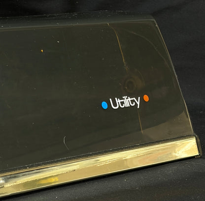 A close-up of a black plastic utility cover with a gold metal edge and the 'Utility' logo printed on it, set against a black fabric background.